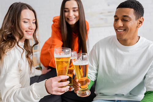 positive women and cheerful african american man toasting glasses of beer in living room