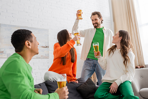 happy bearded man toasting glass of beer near women and looking at african american friend in living room