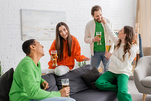 happy multiethnic friends holding glasses of beer while chatting in living room