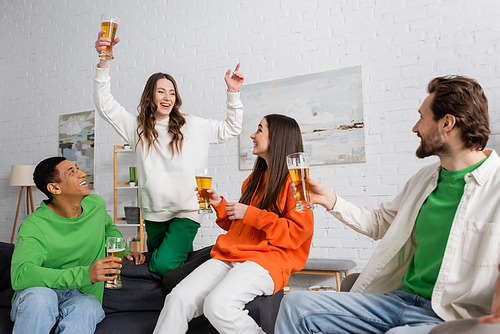 excited woman with glass of beer gesturing near interracial friends in living room