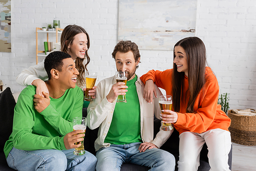 happy interracial group of people looking at bearded friend drinking beer in living room