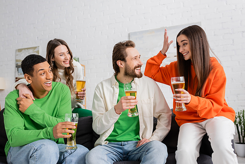 happy interracial people looking at cheerful woman gesturing while holding beer in living room