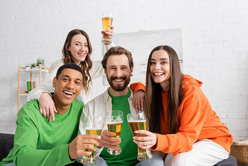 happy interracial group of friends holding glasses of beer in living room