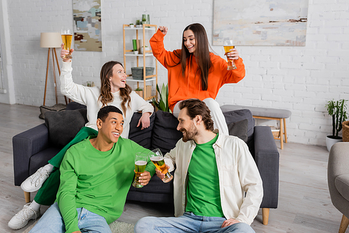 cheerful multicultural men clinking glasses of beer while sitting near happy women in living room