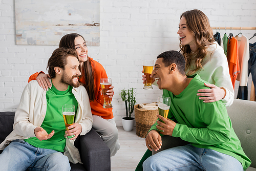 happy women hugging interracial men while holding glasses of beer in living room