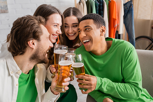 cheerful women and happy interracial men clinking glasses of beer in living room