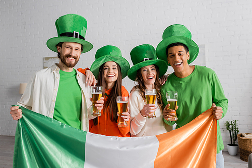 cheerful and multicultural friends in green hats holding glasses of beer and Irish flag while celebrating Saint Patrick Day