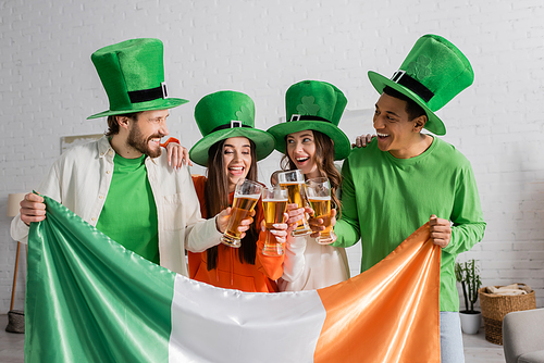 cheerful and multicultural friends in green hats clinking glasses of beer near Irish flag while celebrating Saint Patrick Day