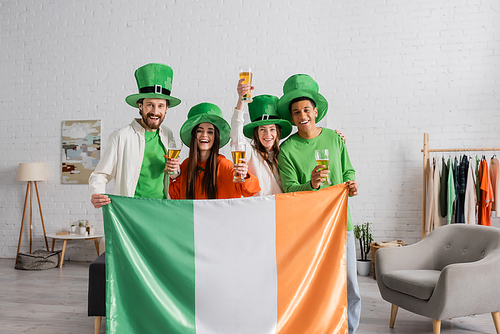 happy and multicultural friends in green hats holding glasses of beer and Irish flag while celebrating Saint Patrick Day