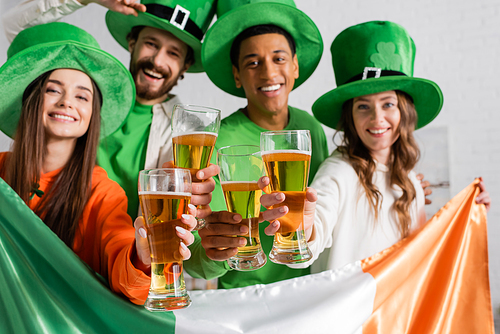 joyful and multicultural friends in green hats holding glasses of beer and Irish flag while celebrating Saint Patrick Day