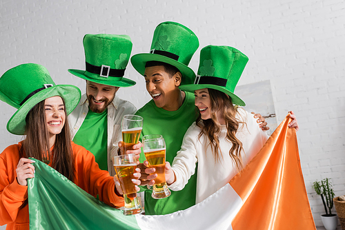 cheerful and interracial friends in green hats holding glasses of beer and Irish flag while celebrating Saint Patrick Day