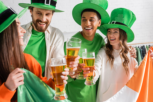 happy and interracial friends in green hats holding glasses of beer and Irish flag while celebrating Saint Patrick Day