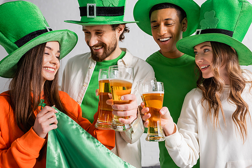 cheerful and interracial friends in green hats clinking glasses of beer near Irish flag while celebrating Saint Patrick Day