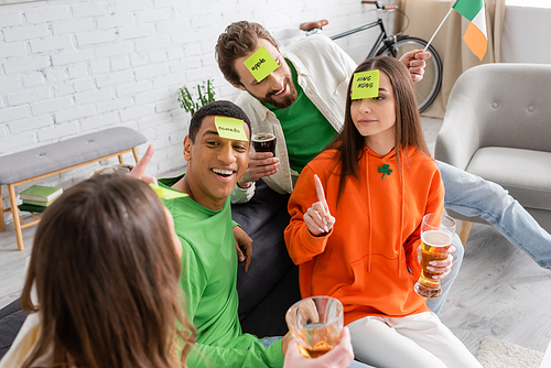 happy interracial friends with sticky notes on foreheads looking at woman while playing guess who game on Saint Patrick Day