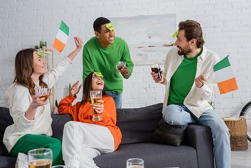 happy interracial friends with sticky notes on foreheads holding drinks and Irish flags while playing guess who game on Saint Patrick Day