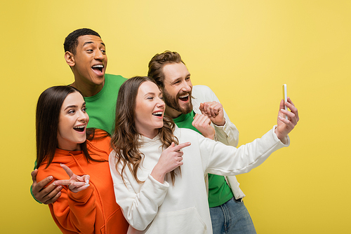 Cheerful interracial friends gesturing and using smartphone isolated on yellow