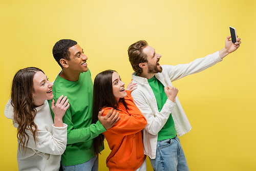 Positive man showing yes gesturing during video call near interracial friends isolated on yellow