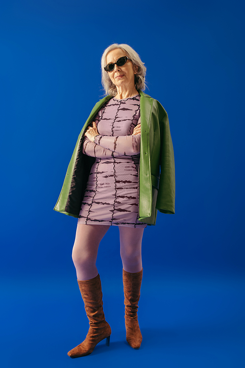 elegant grey haired woman in suede boots and green leather jacket over purple dress standing with crossed arms on blue