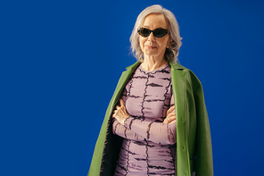 senior woman in stylish outfit and sunglasses standing with crossed arms isolated on blue