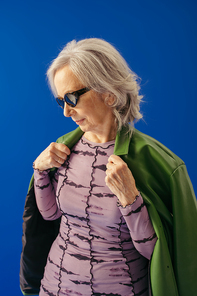 senior woman in stylish sunglasses wearing green leather jacket isolated on blue