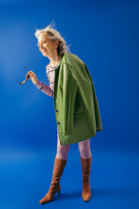 full length of happy senior woman in stylish outfit holding sunglasses on blue