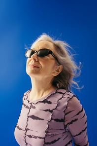 portrait of satisfied senior woman in sunglasses looking away isolated on blue