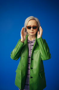 senior woman in green leather jacket adjusting trendy sunglasses isolated on blue