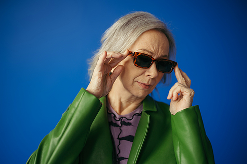 senior woman in green jacket adjusting trendy sunglasses isolated on blue