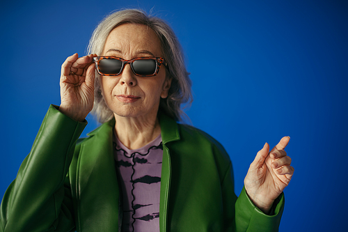 senior woman in green and leather jacket adjusting trendy sunglasses while gesturing isolated on blue