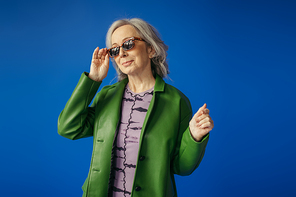 pleased senior woman in green leather jacket adjusting trendy sunglasses isolated on blue