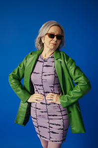 senior woman in green leather jacket and dress standing with hands on hips isolated on blue