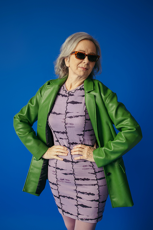 senior woman in green leather jacket and dress standing with hands on hips isolated on blue