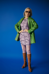 full length of senior woman in green leather jacket and dress standing with hands on hips on blue