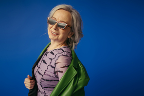cheerful senior woman in sunglasses and green leather jacket smiling isolated on blue