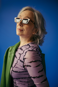 portrait of senior woman in sunglasses standing with green leather jacket isolated on blue