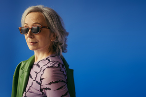 senior woman in sunglasses standing with green leather jacket and looking at camera isolated on blue