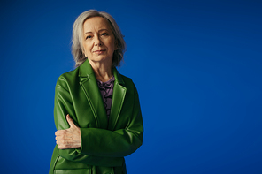 positive grey haired woman in green leather jacket standing with crossed arms isolated on blue