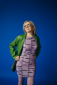positive senior lady in trendy sunglasses and green leather jacket over purple dress standing with hands on hips isolated on blue