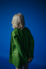 back view of grey haired woman in green leather jacket standing with hands behind back isolated on blue