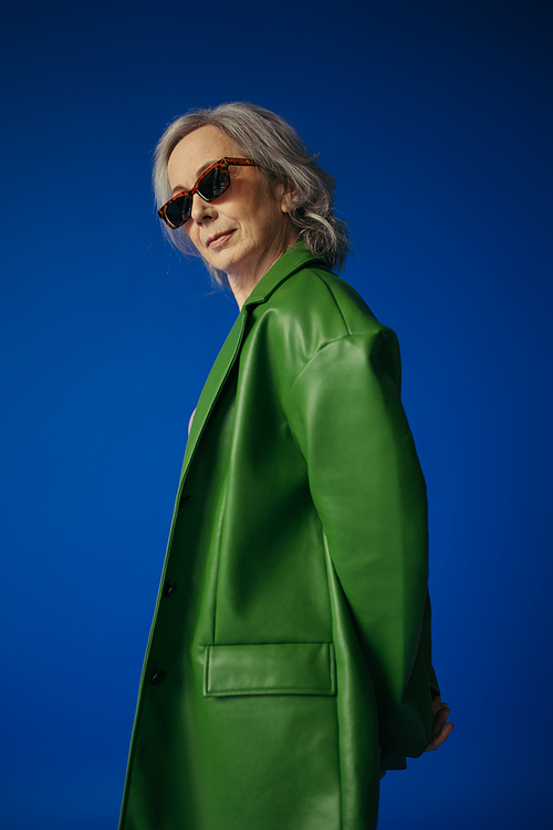 senior model in stylish sunglasses and green leather jacket posing isolated on blue
