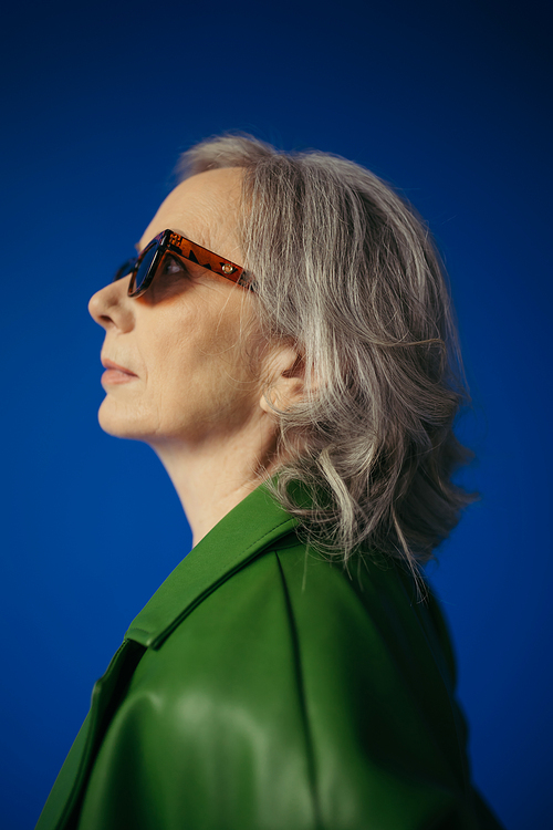 profile of senior grey haired lady in trendy sunglasses and green leather jacket isolated on blue