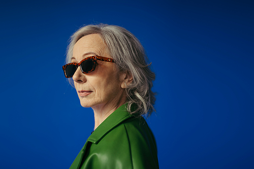 portrait of grey haired woman in green leather jacket and stylish sunglasses isolated on blue