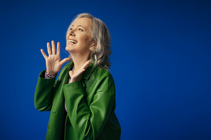 excited grey haired woman in green leather jacket showing wow gesture and looking away isolated on blue