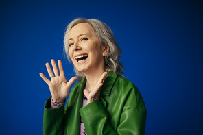 amazed and happy senior woman in green leather jacket showing wow gesture and looking at camera isolated on blue