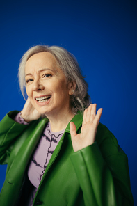 happy grey haired woman in stylish casual clothes waving hand while looking at camera isolated on blue