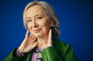 portrait of positive and stylish senior woman posing with hands near neck and smiling at camera isolated on blue