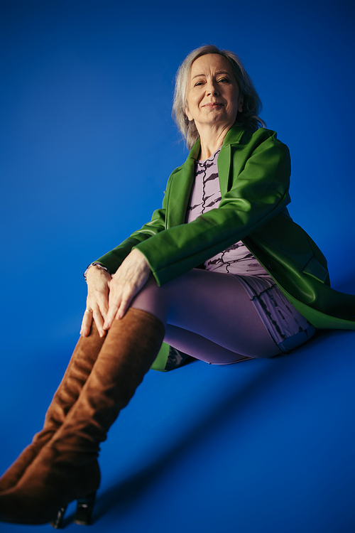 smiling senior model in green leather jacket and brown suede boots sitting on blue background