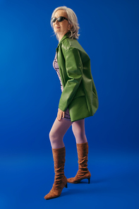 full length of positive senior woman in sunglasses and green leather jacket with brown suede boots on blue background