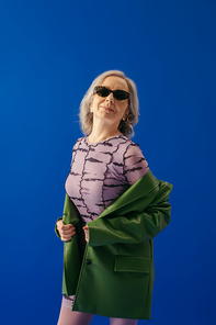 grey haired woman in trendy sunglasses and lilac dress with green leather jacket isolated on blue