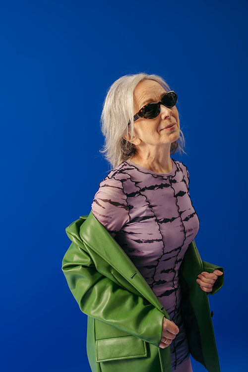 senior woman in sunglasses and green leather jacket over purple dress smiling isolated on blue